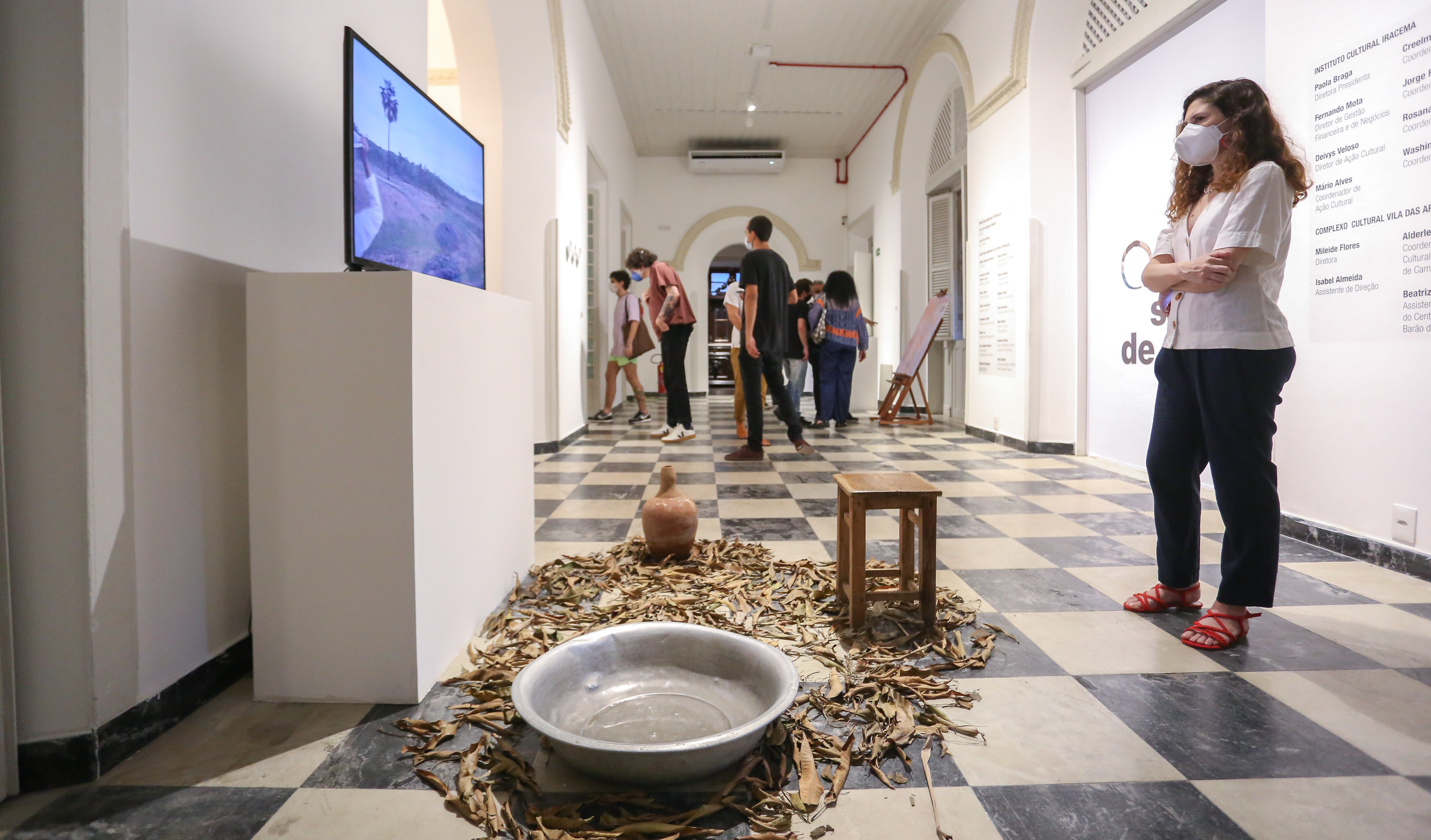 a foto mostra um corredor da casa do barão de camocim com algumas obras do salão de abril, em primeiro plano está uma instalação com uma bacia, uma cabaça e folhas secas espalhadas na frente de uma TV que mostra cenas do sertão